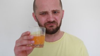 Sarı tişört giymiş kel bir adam şeffaf bir bardak portakal suyu tutuyor, bir yudum alıyor ve sert bir ekşitme ile tepki veriyor, acı bir tadı olduğunu gösteriyor, kapalı, beyaz duvarlı