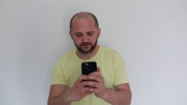 Sarı tişört giyen kel bir adam, beyaz arka planda duran akıllı telefonundan mesaj göndermeye odaklanıyor. Odaklanmış ve elinde cihazla meşgul görünüyor..