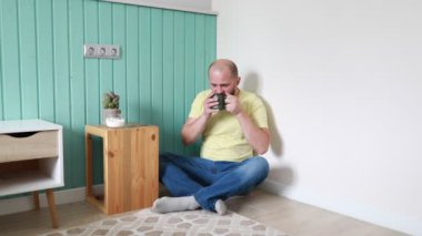 Sarı tişörtlü ve mavi kotlu kel bir adam turkuaz bir duvara yaslanmış yerde rahatça oturuyor. İçten bir ifadeyle, dudaklarına siyah bir kupa getiriyor.