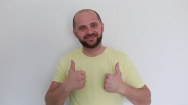 Sarı tişört giyen neşeli kel bir adam düz beyaz bir arka plana karşı duruyor, geniş bir gülümseme, ve coşkuyla iki başparmak kaldırıyor onay ya da başarı işareti olarak.