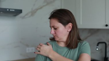 Gündelik yeşil tişörtlü bir kadın, parlak, çağdaş bir mutfakta, aniden rahatsızlık veya acı hisseder. Sabah aktivitelerine ara veriyor gibi görünüyor, elini göğsüne koyuyor.