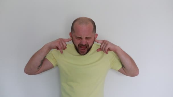 一个穿着黄色T恤的秃头男人站在白色的背景上 用手指捂住耳朵 脸上挂着鬼鬼祟祟的表情 暗示着他在试图挡住一声巨响或表现出一种奇怪的声音 — 图库视频影像