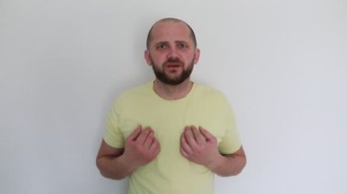 Soluk sarı gömlek giyen sakallı bir adam düz beyaz bir arka plana karşı durur, omuz silker ve açık avuçlarını gösterir evrensel bir kafa karışıklığı veya belirsizlik jesti.