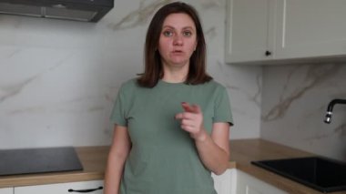 Günlük tişörtlü bir kadın çağdaş bir mutfakta duruyor, parmaklarıyla bir numarayı işaret ediyor, belki de bir sunum veya konuşma sırasında önemli bir noktayı vurguluyor..
