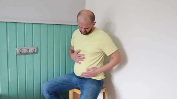 一个穿着黄色T恤的男人不自在地笑着 紧紧抓住他的胃 坐在蓝色绿色镶板墙上的木制凳子上 他脸上的痛苦表情表明他可能是 — 图库视频影像