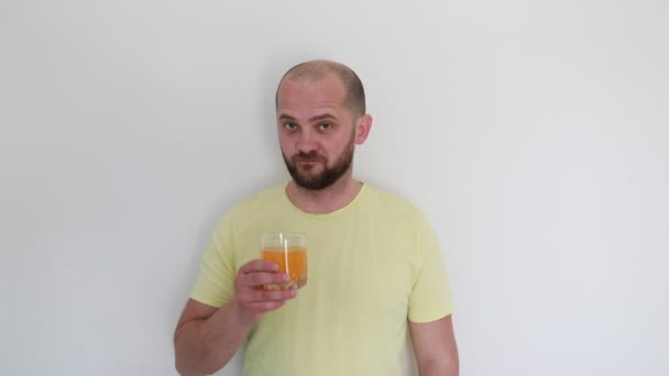 明るい黄色のTシャツを着た男性が オレンジジュースをグラスにした白い壁に向かって立っています 彼は一口を飲んで 思慮深い表現で味を節約し 評価しているように見えます — ストック動画