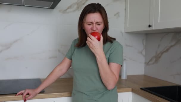 女性は明るくコンテンポラリーなキッチンに立って 鮮やかな赤いリンゴを噛みます 彼女は シンプルで健康的な生活の瞬間を反映して 清潔で明るいスペースで満足して表示されます — ストック動画