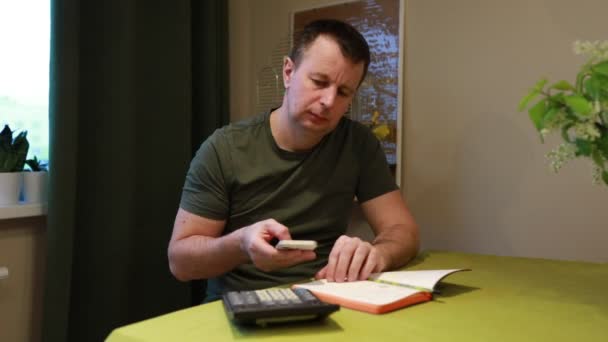 男はテーブルに座って 深く集中して 彼の目の前に本と彼の側の計算機が開いている 彼は勉強したり 必要とされる仕事に取り組んでいるようです — ストック動画