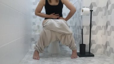 Banyoda tuvalette oturan bir kadını gösteren bir video. Kadın tuvaleti kullanıyor ve görünüşe göre özel bir yerde. Odak noktası sadece tuvalette oturma eylemi..