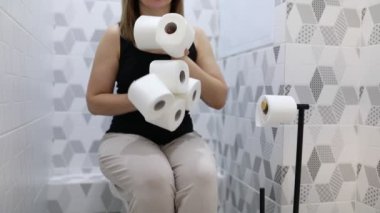 Bir kadın, elinde iki rulo tuvalet kağıdı tutarken klozet kapağında otururken görülüyor. Banyo ortamında içeride gibi görünüyor..