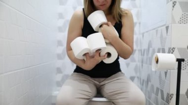 Tuvalette oturan bir kadın, elinde üç rulo tuvalet kağıdı tutuyor. Bir banyo ortamında, tipik bir banyo aktivitesiyle meşgul gibi görünüyor..