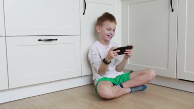 Genç bir çocuk, el bilgisayarı konsolunda oyun oynamaya dalmış, parlak mutfak zemininde bacak bacak üstüne atarak oturuyor. Odaklanmış ifadesi ve günlük kıyafetleri evde rahat bir günü yansıtıyor..
