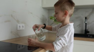 Genç bir çocuk mutfak tezgahında duruyor ve temiz bir sürahiden bardağa su döküyor. Modern mutfak seti doğal ışıkla ışıl ışıl aydınlatılıyor.