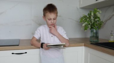Mutfakta elinde tabletle duran genç bir çocuk var. Gördüklerine dalmış gibi görünüyor, belki bir oyun ya da eğitici içerik. Mutfak çevresi görülebilir durumda.