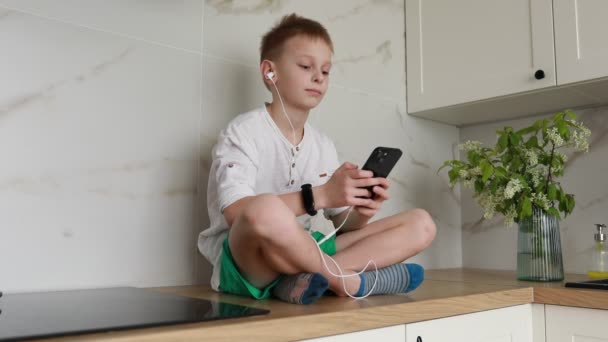 一个小男孩舒服地坐在厨房的台面上 全神贯注地听着音乐 用白色耳塞连接着他的手持智能手机 房间里充满了自然光 凸显了休闲的气氛 — 图库视频影像