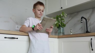 Genç bir çocuk mutfakta elinde bir tablet tutarken görülüyor. Ekrana odaklanmış görünüyor, muhtemelen video izliyor ya da oyun oynuyor. Mutfak temiz, tezgahları var ve...