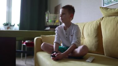 Genç bir çocuk bir kanepede sakince oturuyor, bir video oyunu oynamaya kendini kaptırmış. Oyuna odaklanmış, bir kumanda tutuyor ve ekrandaki harekete tepki veriyor..