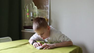 Odaklanmış genç bir çocuk, sarı bir masa örtüsüyle kaplı bir yemek masasında oturur, özenle bir kağıt parçasına çizim yapar ve çalışma alanına yumuşak bir ışık tutar.