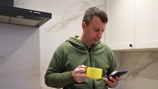 男はキッチンに立って 注意深く携帯電話の画面を見ている 彼がデバイスと相互作用するように集中し 従事しているように見えます おそらくそれで何かを読んだり 見たりします — ストック動画