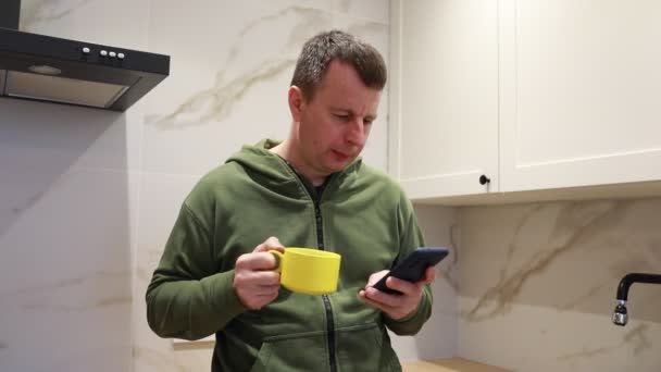 Mężczyzna Stoi Kuchni Trzymając Kubek Jednej Ręce Komórkę Drugiej Wygląda — Wideo stockowe