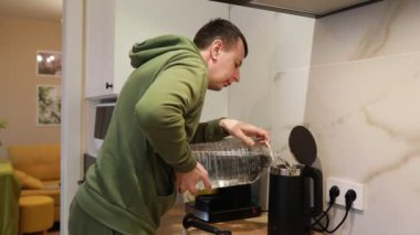 Odaklanmış bir adam, iyi aydınlatılmış, çağdaş bir mutfakta büyük bir su şişesinden elektrikli su ısıtıcısını dolduruyor. Akşam vakti gibi görünüyor ve muhtemelen sıcak bir içecek için su kaynatmaya hazırlanıyor.