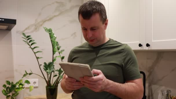 一个专心致志的男人站在灯火通明的厨房里 专心致志地使用平板电脑 他穿着宽松的绿色衬衫 似乎沉迷于屏幕上的内容 可能会浏览食谱或阅读新闻 — 图库视频影像
