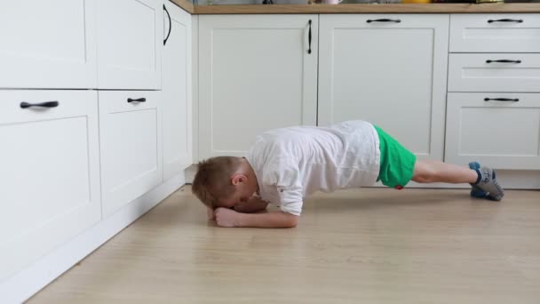 一名身穿绿色衬衫和短裤的小男孩在厨房的白色地板上热切地做俯卧撑 展示出一种健康的生活方式 — 图库视频影像