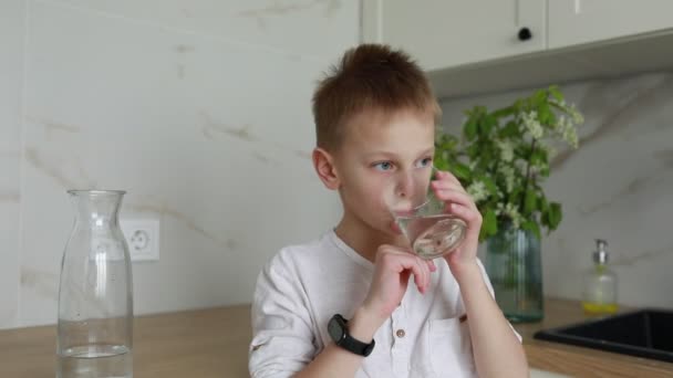 白いシャツを着た少年が 明るく現代的なキッチンに立っている 水分補給され リフレッシュされた水から 清潔で新鮮な水を飲む — ストック動画
