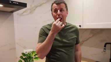 Bir adam iyi ışıklandırılmış, modern bir mutfakta duruyor, tavadan yiyecek örnekleri alıyor. Yemeğin tadını alırken dalgın görünüyor. Muhtemelen tadını değerlendiriyor ya da bir sonraki hamlesinin ne olacağına karar veriyor..