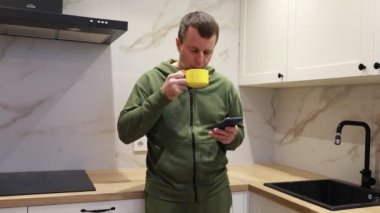 Bir adam mutfakta cep telefonu tutarken ve dikkatle ekrana bakarken görülüyor. Cihaza odaklanmış ve meşgul görünüyor, muhtemelen bir mesaj okuyor, sosyal medyayı kontrol ediyor, ya da