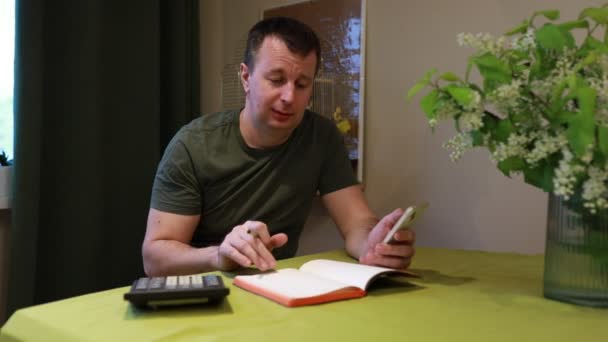 Mężczyzna Siedzi Przy Domowym Stole Pisząc Notatki Notatniku Długopisem Sporadycznie — Wideo stockowe