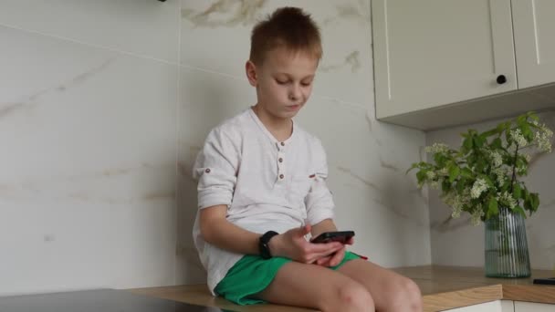 一个小男孩坐在柜台上 手里拿着一部手机 他专注于设备屏幕 拍击和滑动 这个男孩似乎专注于数字内容 对数字内容视而不见 — 图库视频影像