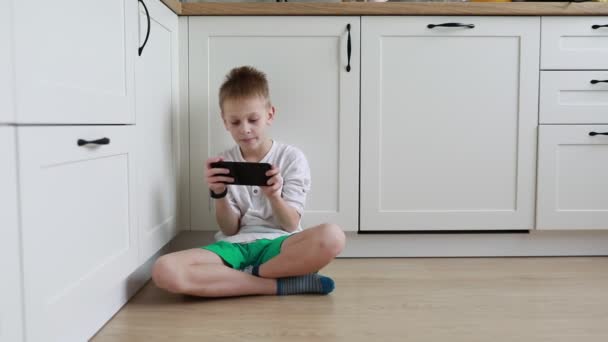 一个专心致志的小男孩两腿交叉坐在厨房的地板上 全神贯注地在手持式控制台上玩游戏 身后的房间里闪烁着阳光 — 图库视频影像