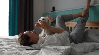 Güneş ışığı odayı hafifçe aydınlatırken, neşeli bir evcil hayvan sahibi, rahat bir yatakta, küçük beyaz köpekleriyle sevgi dolu oyun zamanını paylaşıyor, bu da aralarında yakın bir bağ olduğunu gösteriyor..