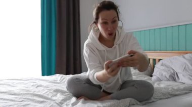 Bir kadın rahat bir şekilde yatağında bağdaş kurmuş, rahat bir pijama ve kapüşonlu bir kazak giyiyor. Yüzünde bir gülümsemeyle, akıllı telefonuyla bir oyun oynamaya dalmıştı, yumuşak sabah ışığı.