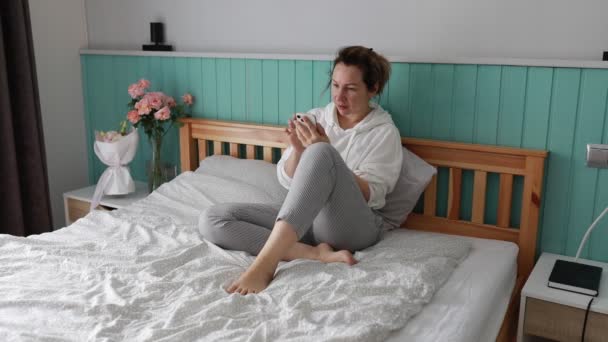 一个女人舒舒服服地坐在床上 双腿交叉 沉浸在智能手机里 卧室里有一种宁静的氛围 配有木制的床头板 充满了鲜花 — 图库视频影像