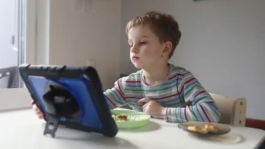 Genç bir çocuk mutfak masasında oturur, kahvaltısını yaparken tablet ekranına derinden odaklanır. Sabah ışığı odayı hafifçe aydınlatıyor, çocuğun ifadesini vurguluyor.