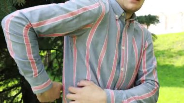 Genç bir adam yemyeşil bir parkta dimdik ayakta duruyor, duruşu güven saçıyor. Parlak güneş ışığı ağaçların arasından süzülüp sahneyi aydınlatıyor. Çizgili gömleği ve günlük tarzı...