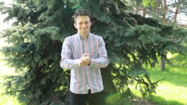 Ağacın önünde duran bir adam iyi niyet göstergesi gösterir. Gülümsüyor ve park ortamında kameraya bakıyor..