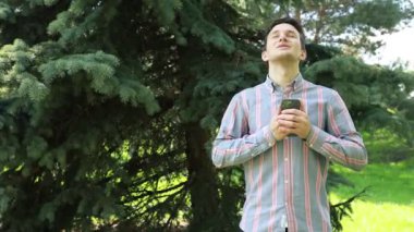 Çizgili gömlekli genç bir adam, akıllı telefonuna dalmış, yemyeşil bir parktaki bir ağacın gölgesinde duruyor. Baharın parlak öğleden sonrası güneşi, huzurlu yeşilliğin üzerine yumuşak bir ışık saçar.