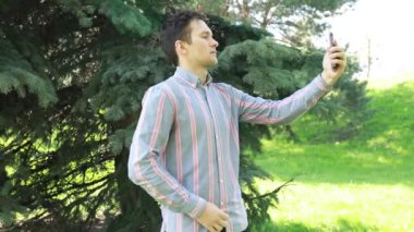 Bir adam bir ağacın önünde elinde bir cep telefonu tutarken görülüyor. Cihaz ekranına odaklanmış gibi görünüyor, belki fotoğraf çekiyor ya da mesajlaşıyor. Arka plan tipik bir açık hava göstergesi