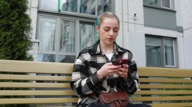 Genç bir kadın akıllı telefonuyla meşgul, bankta otururken dikkatle mesajlaşıyor. Arka planda modern binaların olduğu bir şehir parkı. Bu da bir bağlantı anının olduğunu gösteriyor.