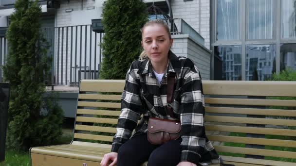 平らなシャツとジーンズの若い女性は 思慮深い表情で木の公園のベンチに座り おそらく誰かを待ったり 孤独の瞬間を楽しんだりします 設定は都市であるように見えます — ストック動画