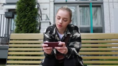 Odaklanmış genç bir kadın, güneşli bir park bankında oturuyor, akıllı telefonuyla meşgul oluyor, konsantrasyon bakışlarıyla içeriği arasında kayıyor. Günlük kıyafetleri, zaman geçirdikçe rahatladığını gösteriyor.
