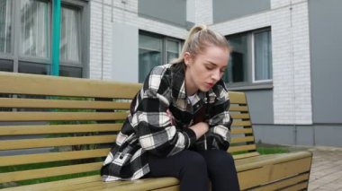 Genç bir kadın açık hava bankında oturuyor, karnını tutuyor ve rahatsızlık ya da acı belirtileri gösteriyor. Ani mide ağrısıyla boğuşuyor gibi görünüyor.