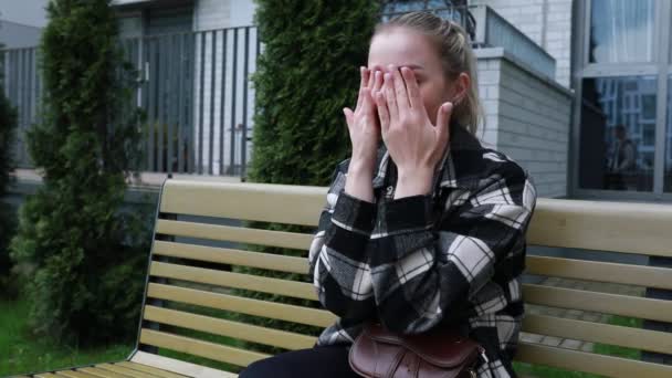 一位年轻女子穿着一件休闲格子呢夹克 坐在外面的木制公园长椅上 她显然心烦意乱 双手捂住脸 露出痛苦或悲伤的表情 — 图库视频影像