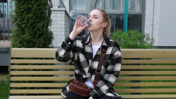 若い女性が木製の公園のベンチに座り 透明なプラスチック製の水のボトルからさわやかな飲み物を取ります チェッカーのジャケットで気軽に服を着た彼女は 都市公園で休憩を楽しんでいるようです — ストック動画