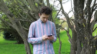 Bir adam bir ağacın önünde duruyor, cep telefonu ekranına dikkatle bakıyor. Cihaza odaklanmış görünüyor, muhtemelen mesajları kontrol ediyor ya da internete bakıyor. Ağaç ona arka plan sağlar.
