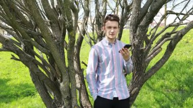 Gündelik gömlekli genç bir adam yapraksız ağaçların arasında duruyor, açık bir bahar gününün parlak güneşinin altında akıllı telefonuyla kitap okuyor ya da daktilo yazıyor..