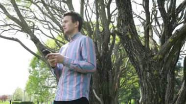 Bir adam cep telefonunu kullanırken bir ağacın önünde dikilirken görülüyor. Açık hava ortamında onunla etkileşime girerken cihaza odaklanmış gibi görünüyor..
